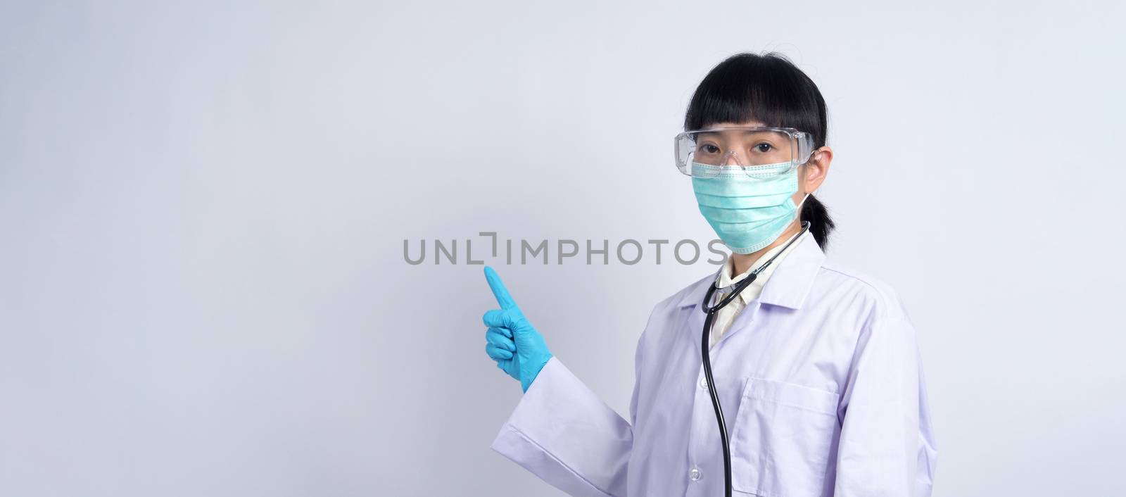 Asian woman doctor pointing finger out to copy space beside her and she wearing white color suit and medical mask and blue rubber gloves to prevent virus pandemic and stethoscope on neck.