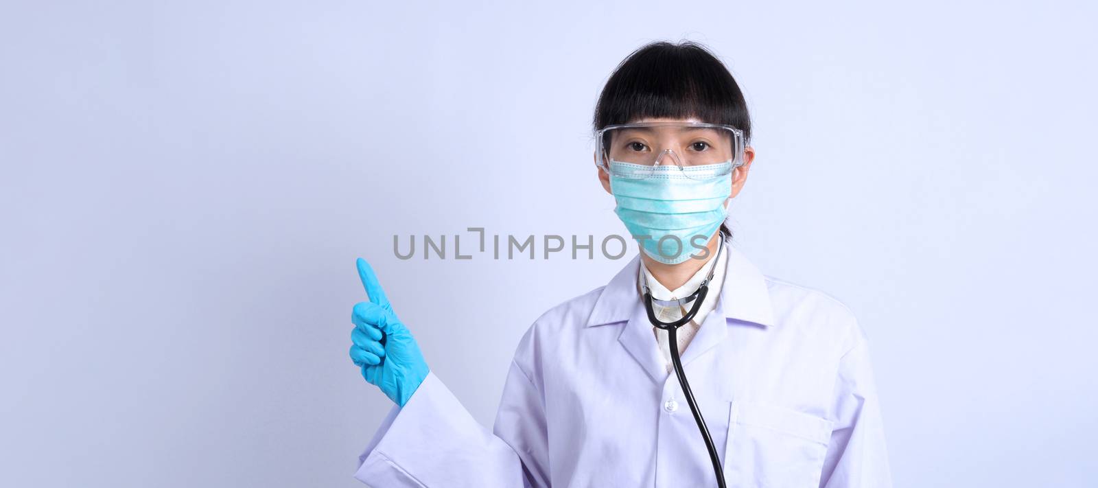 Asian woman doctor pointing finger out to copy space beside her and she wearing white color suit and medical mask and blue rubber gloves to prevent virus pandemic and stethoscope on neck.