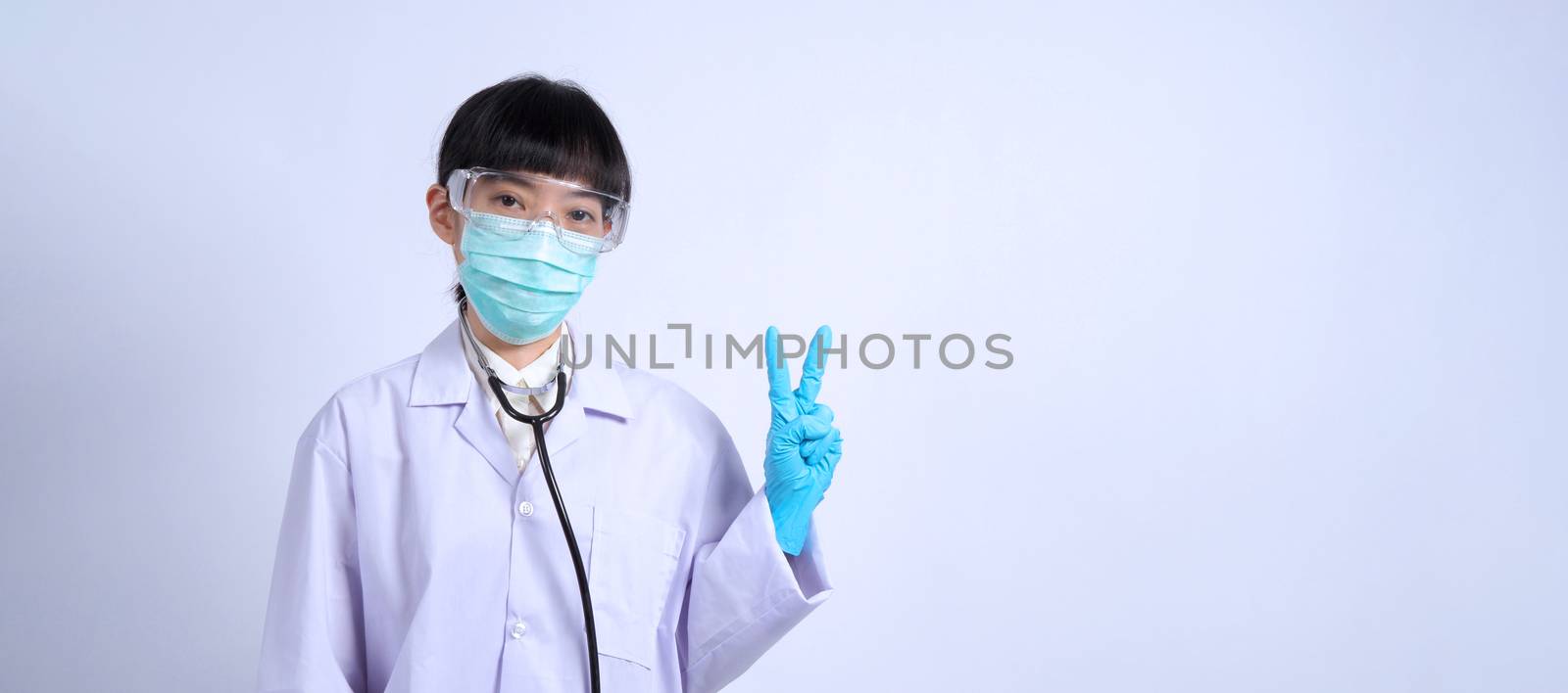 Asian woman doctor pointing finger out to copy space beside her and she wearing white color suit and medical mask and blue rubber gloves to prevent virus pandemic and stethoscope on neck.