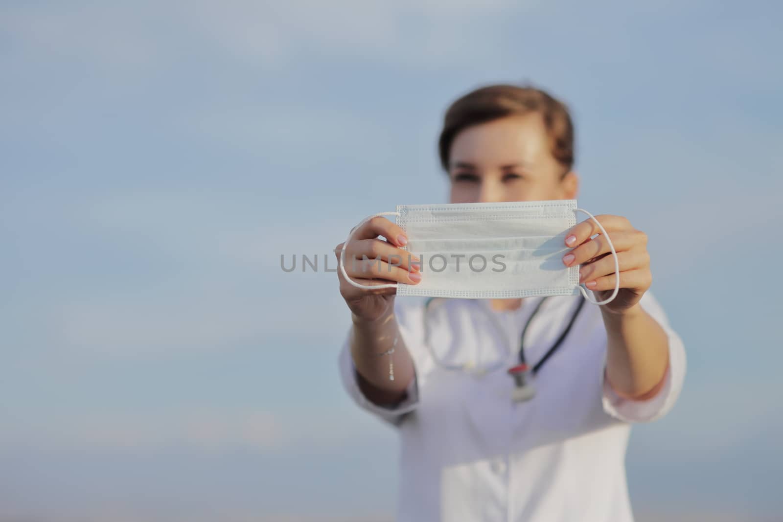 Young woman doctor or nurse showing medical face mask close up. Blue sky with clouds. Prevention Covid-19 healthcare concept. Stethoscope over the neck. Female, girl.