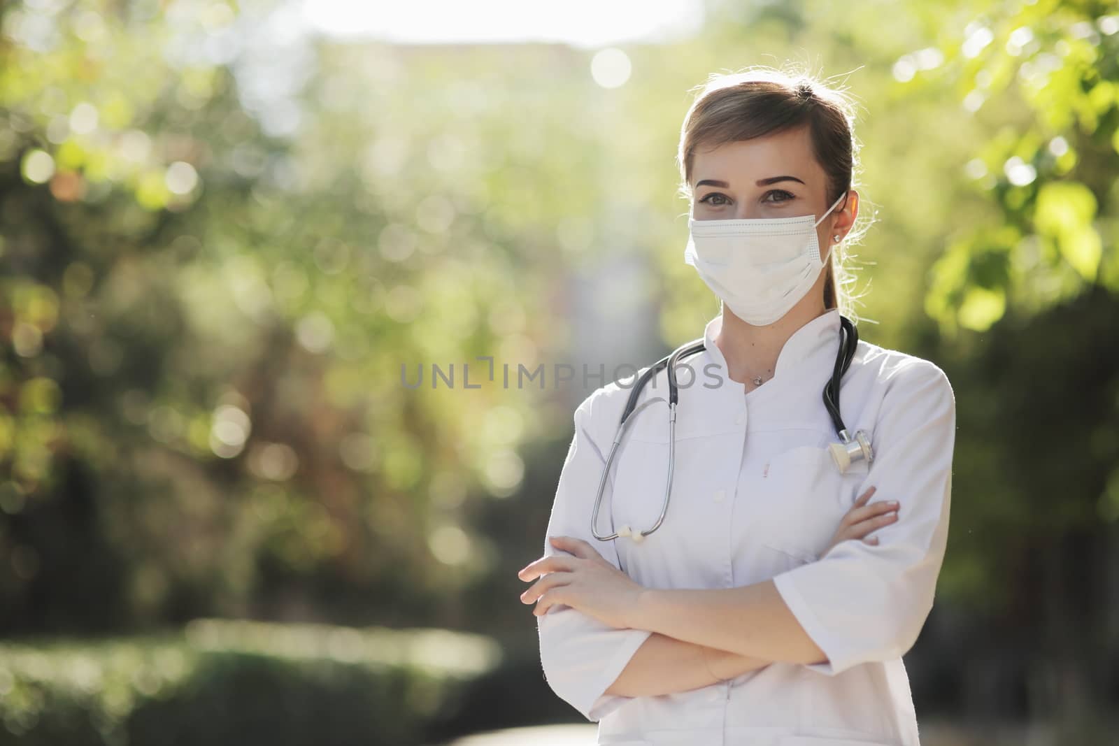 Woman doctor or nurse in a face protective mask in the park among the trees. Safety measures against the coronavirus. Prevention Covid-19 healthcare concept. Stethoscope over the neck. Woman, girl.