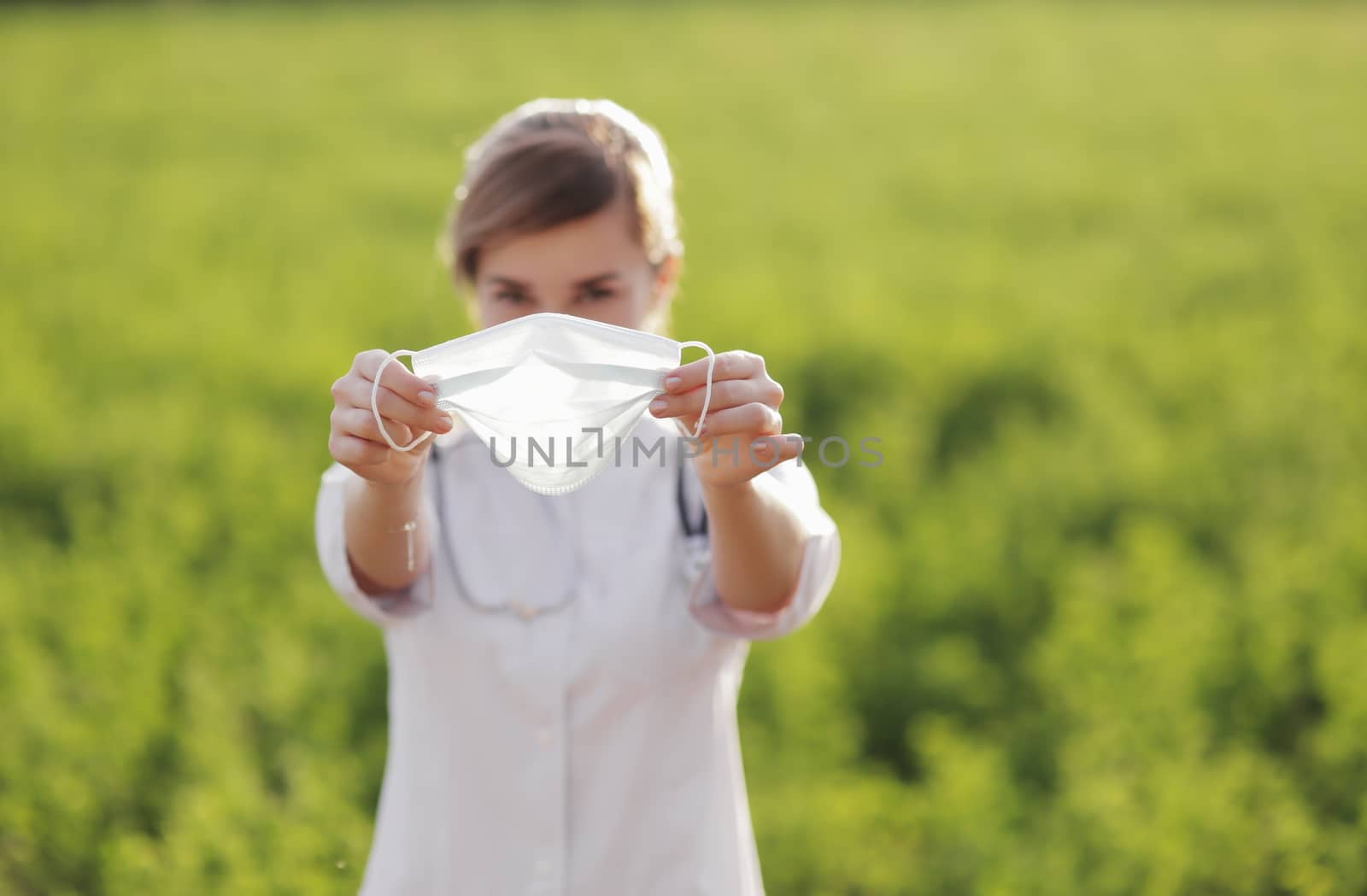 Young woman doctor or nurse showing medical face mask close up. On green grass background. Prevention Covid-19 healthcare concept. Stethoscope over the neck. Female, girl.