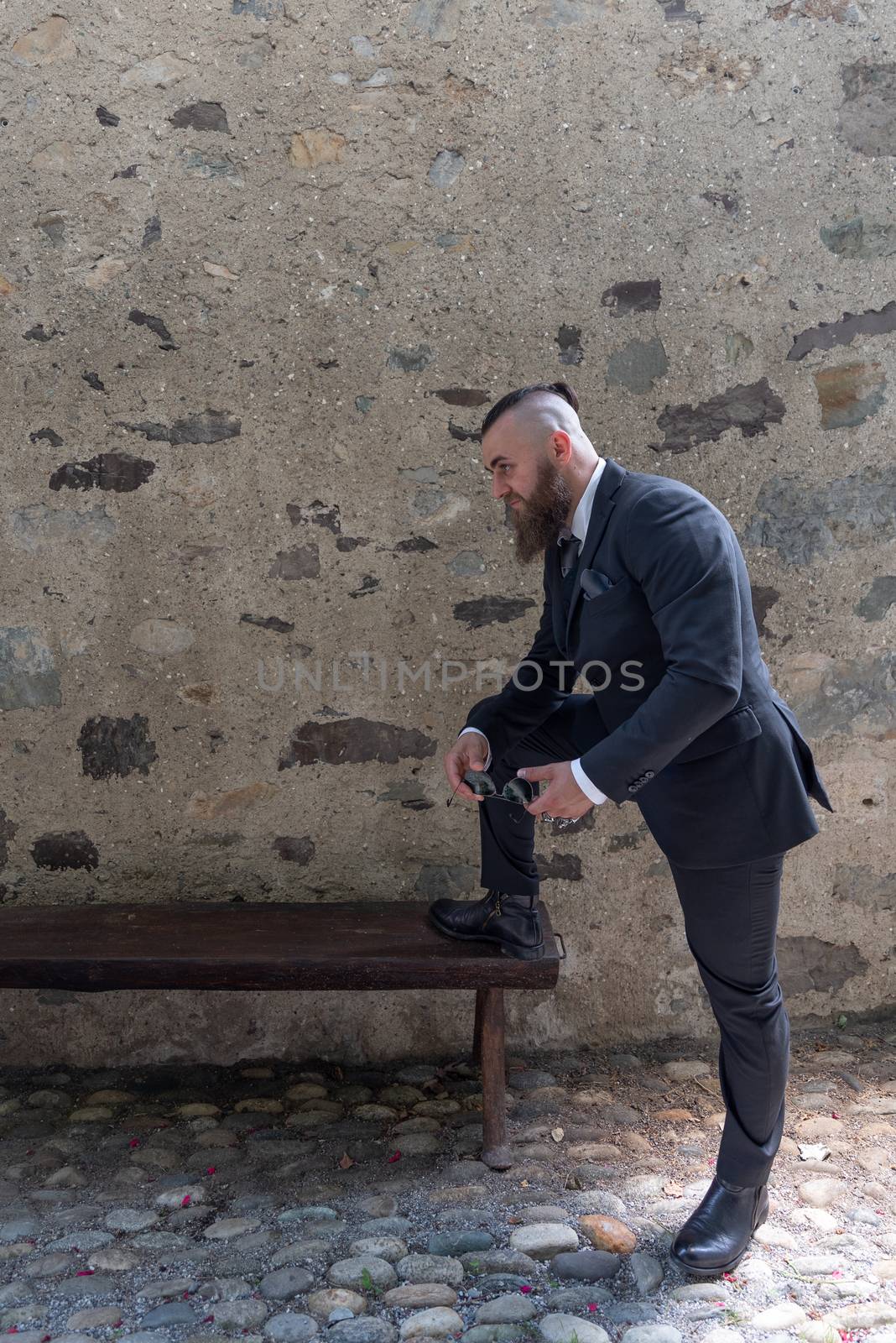 Man in elegant clothes rests one foot on a wooden bench in a small village
