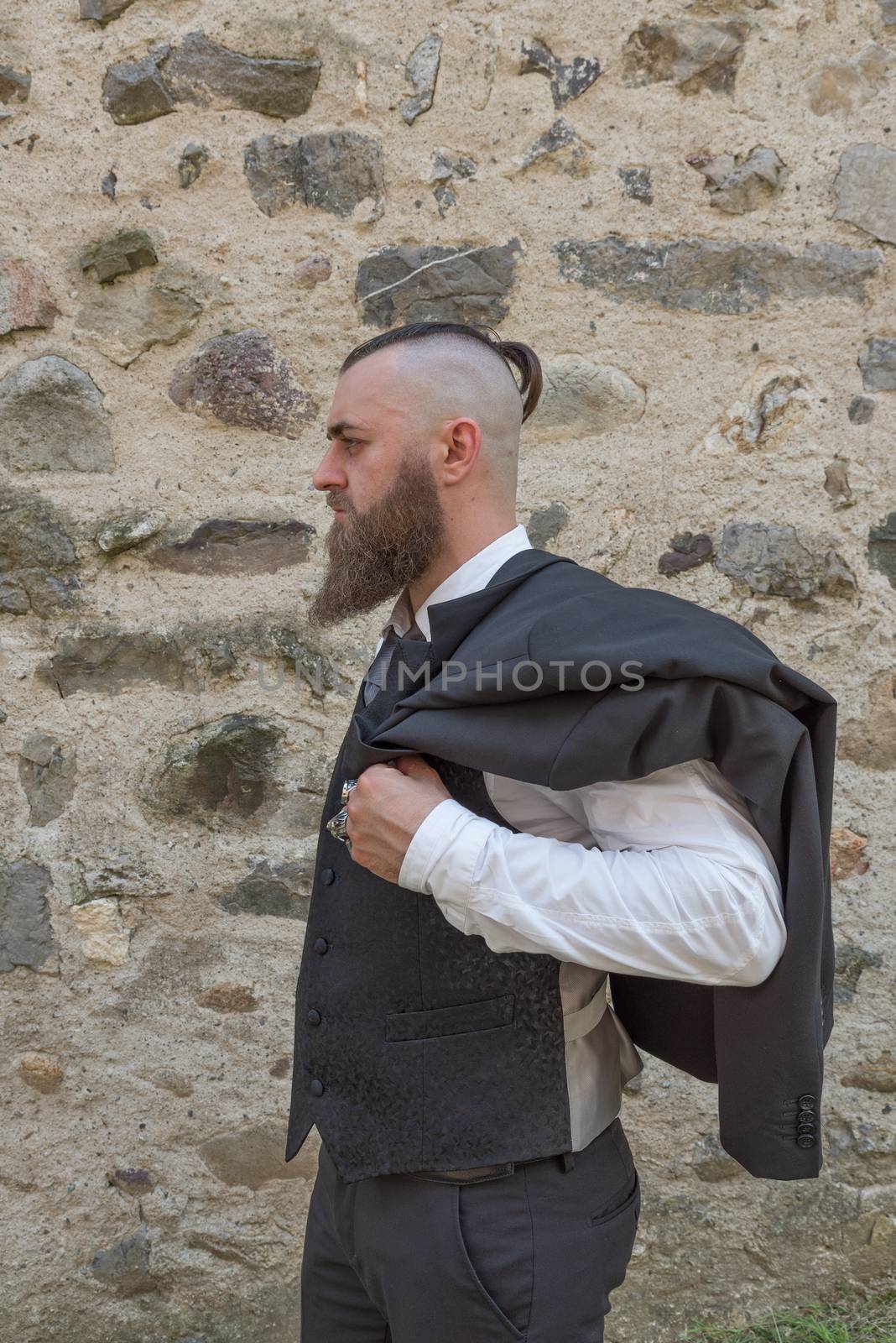 Man with long beard wears a dark elegant suit posing in front of a stone wall