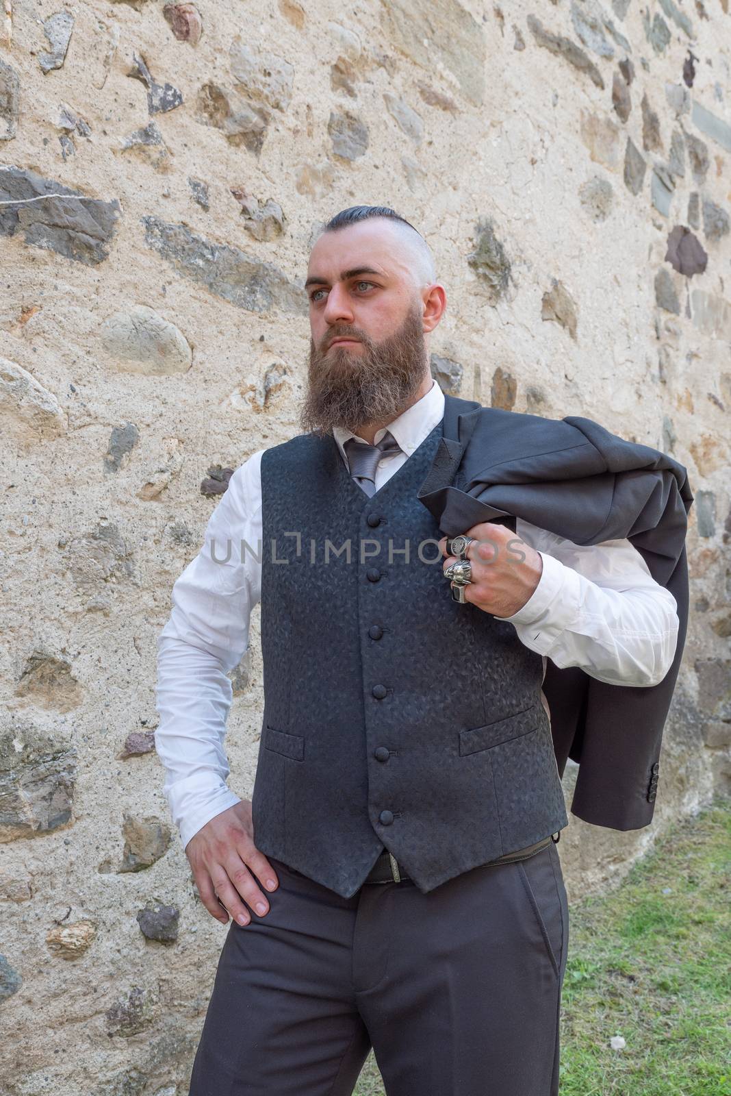 Man with long beard wears a dark elegant suit posing in front of a stone wall