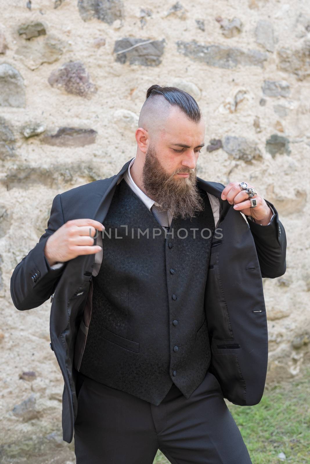 Man with long beard wears a dark elegant suit posing in front of a stone wall