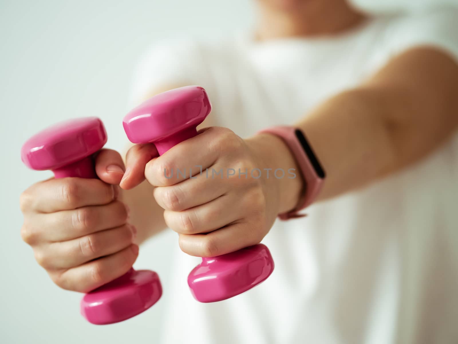 Unrecognizable young woman in white shirt with pink wearable device and bright pink colored dumb-bell in hands. Focus on barbells in female hands