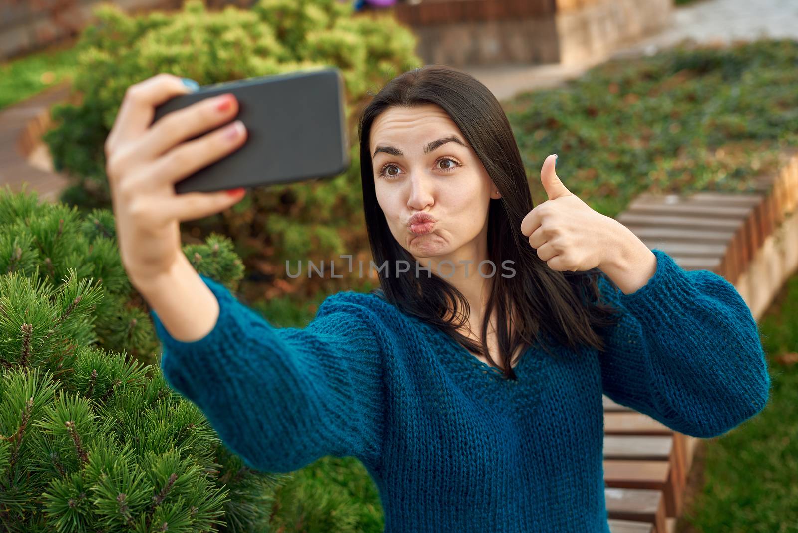 Pretty female student sending kiss and showing thumb up while making photo of herself outside of a green cosy frontage