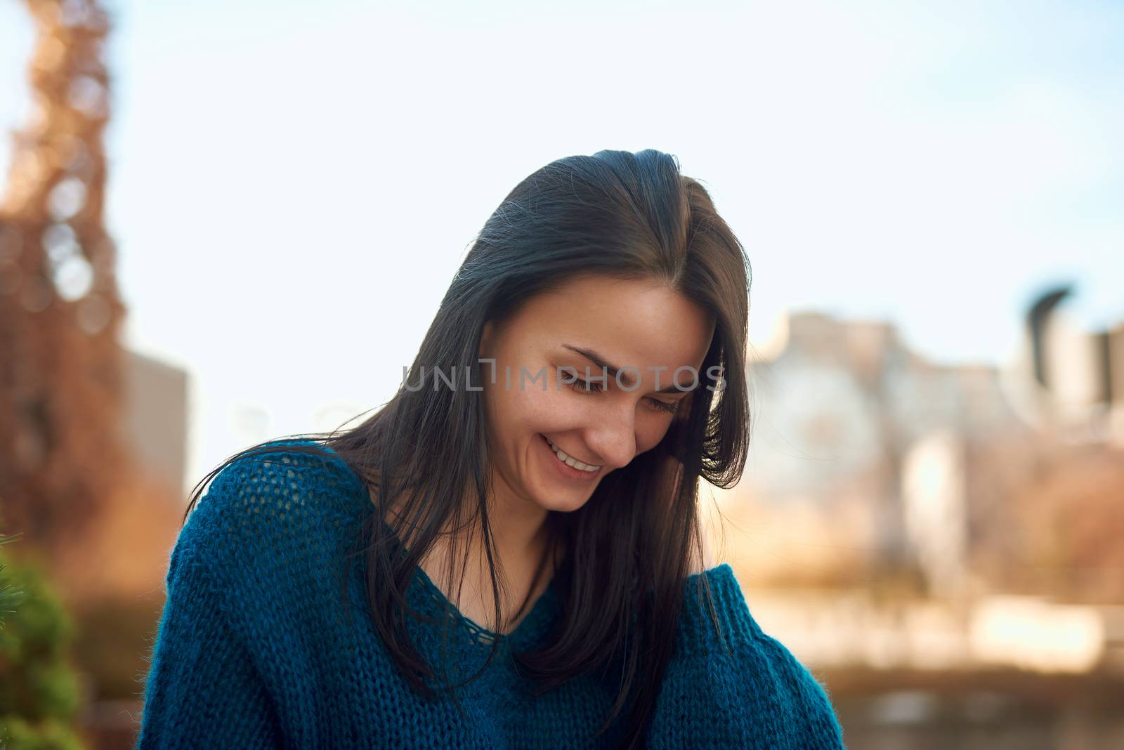 Young beautiful woman looking down with happy smile while tidying up her hair with hand on defocused background of city street