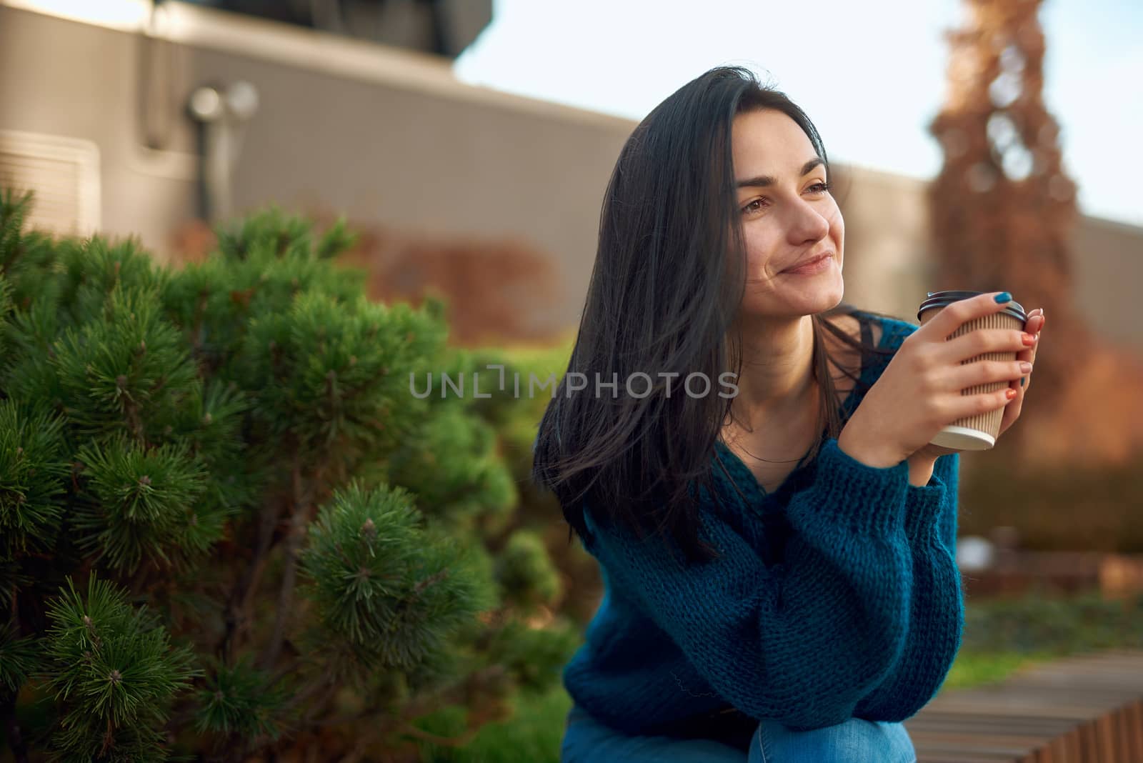 Romantic young lady with lovely smile and far away look holding cup of coffee while sitting on bench in square