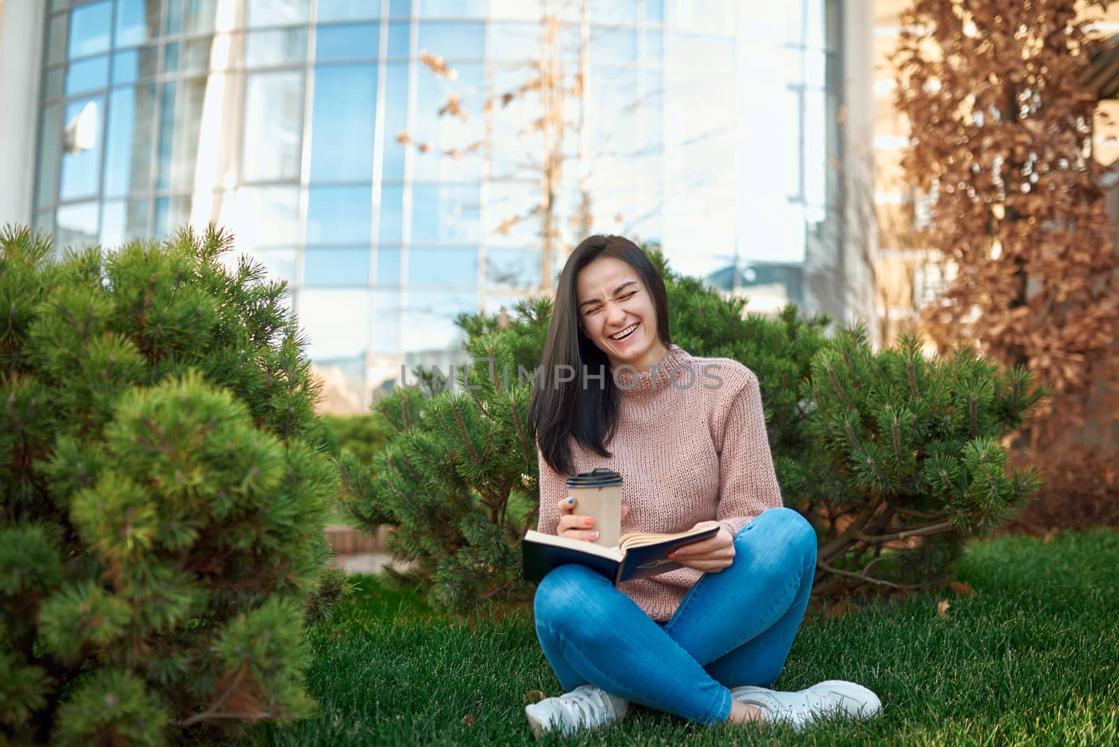 Nice-looking female student laughing enjoying funny book on a cosy green lawn nearby a tall city tower