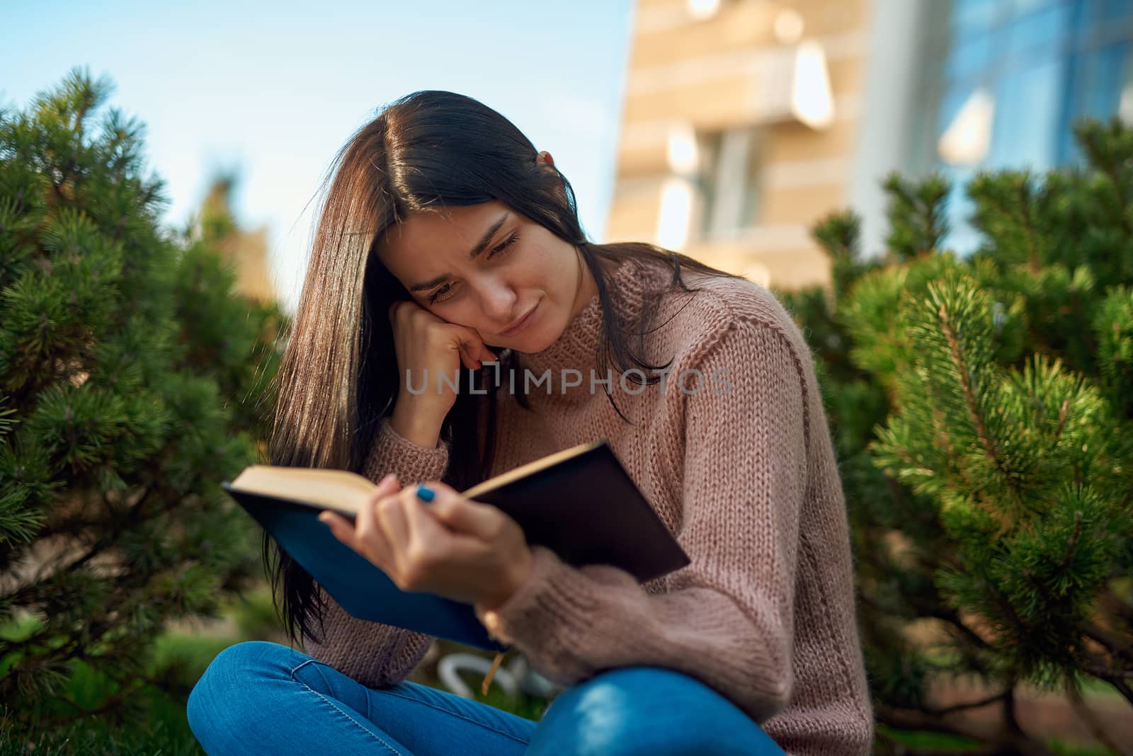 Thoughtful concentrated young woman sitting in a cosy pose on a lawn among tiny conifers, leaning her cheek on hand while reading a serious book