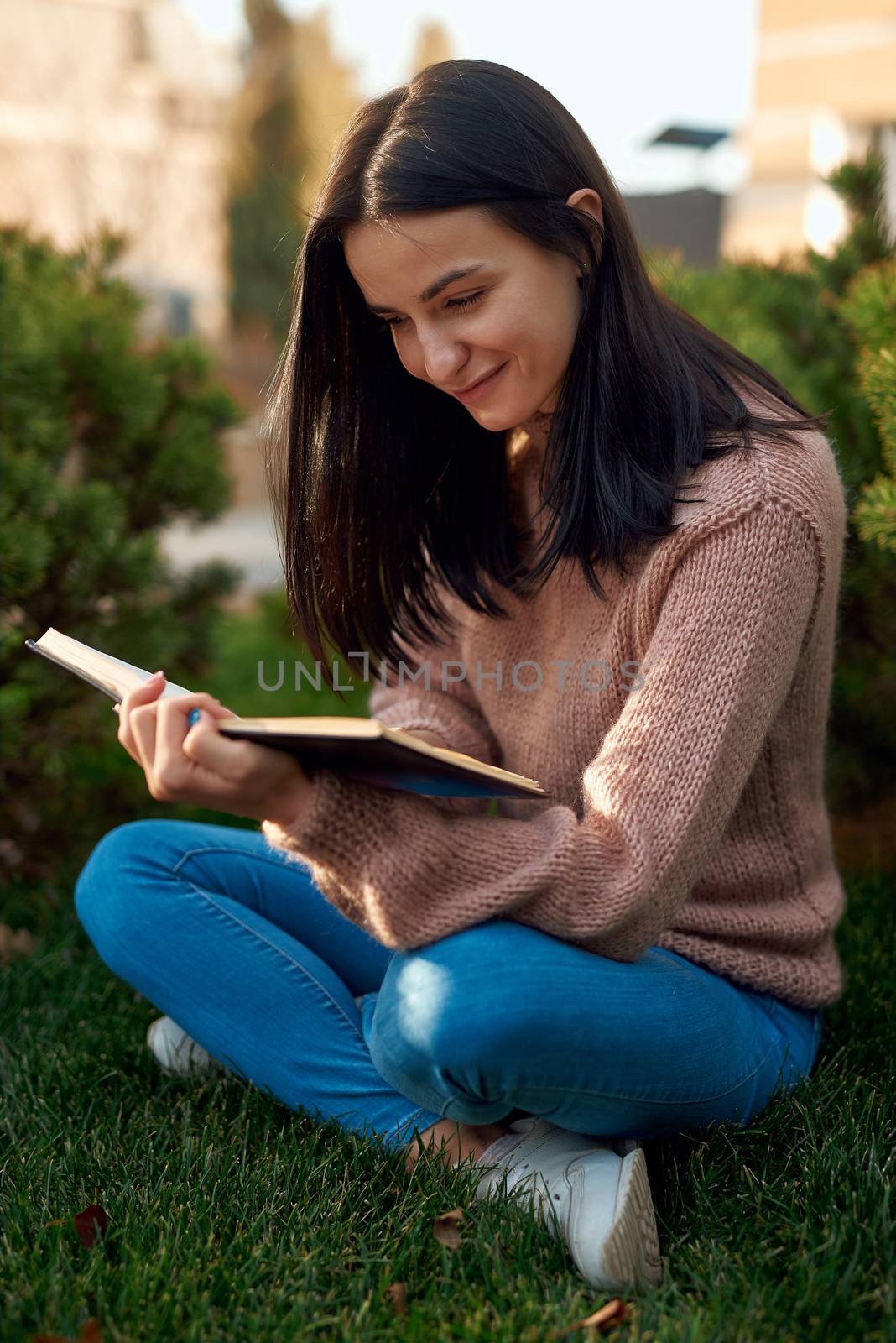 Tender female brunette in casual clothing posing on a green lawn with her leggs crossed with a good novel