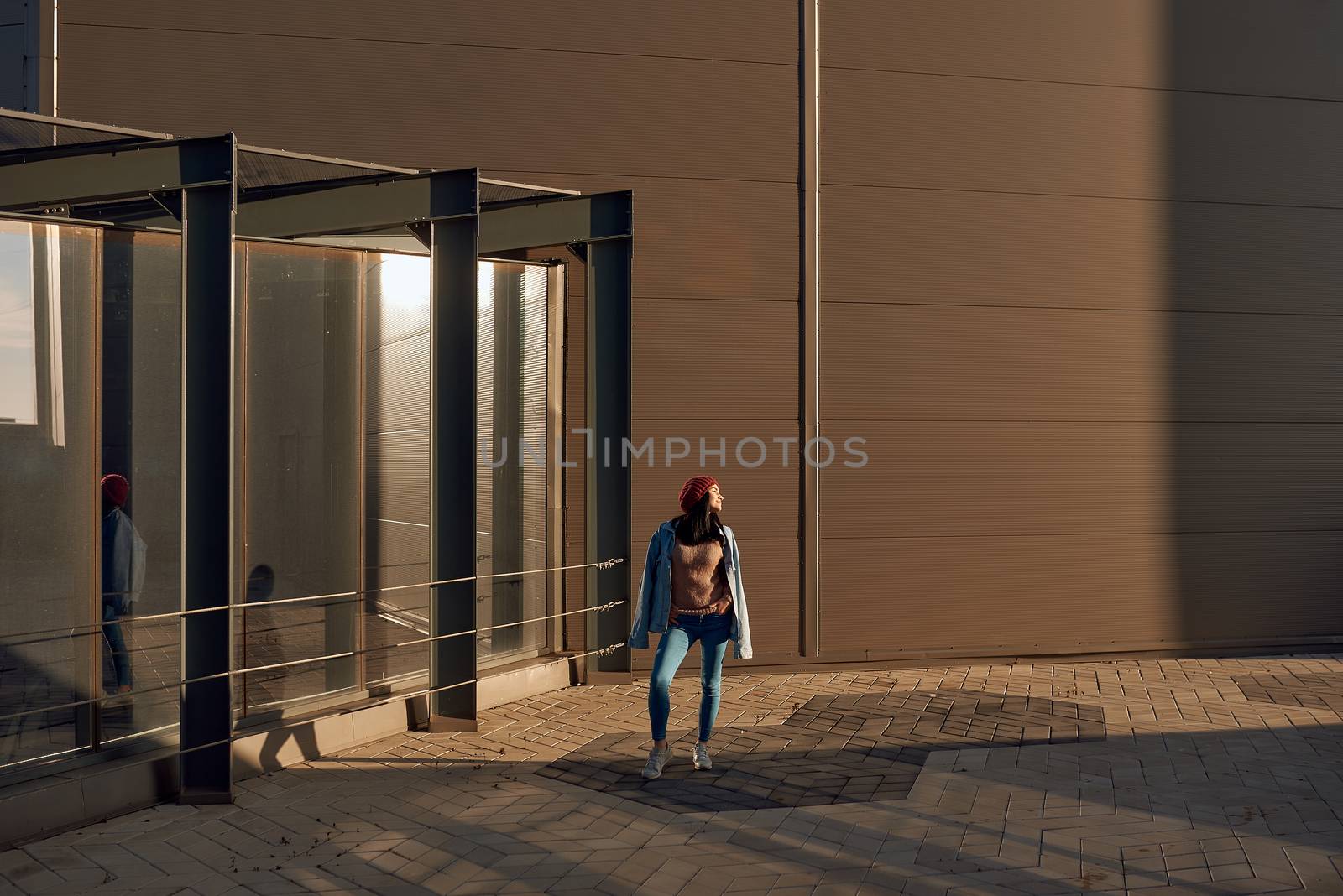 Beautiful young woman brunette in trendy jeans outfit standing close to a modern city building and looking aside