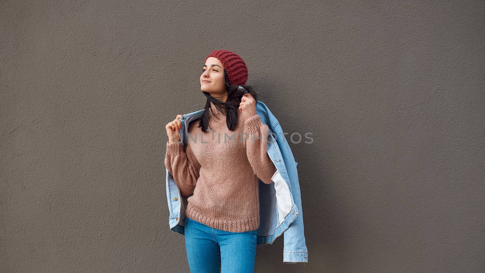 Pleasant young lady in stylish attire wrapping herself up in denim coat while looking aside near a large wall