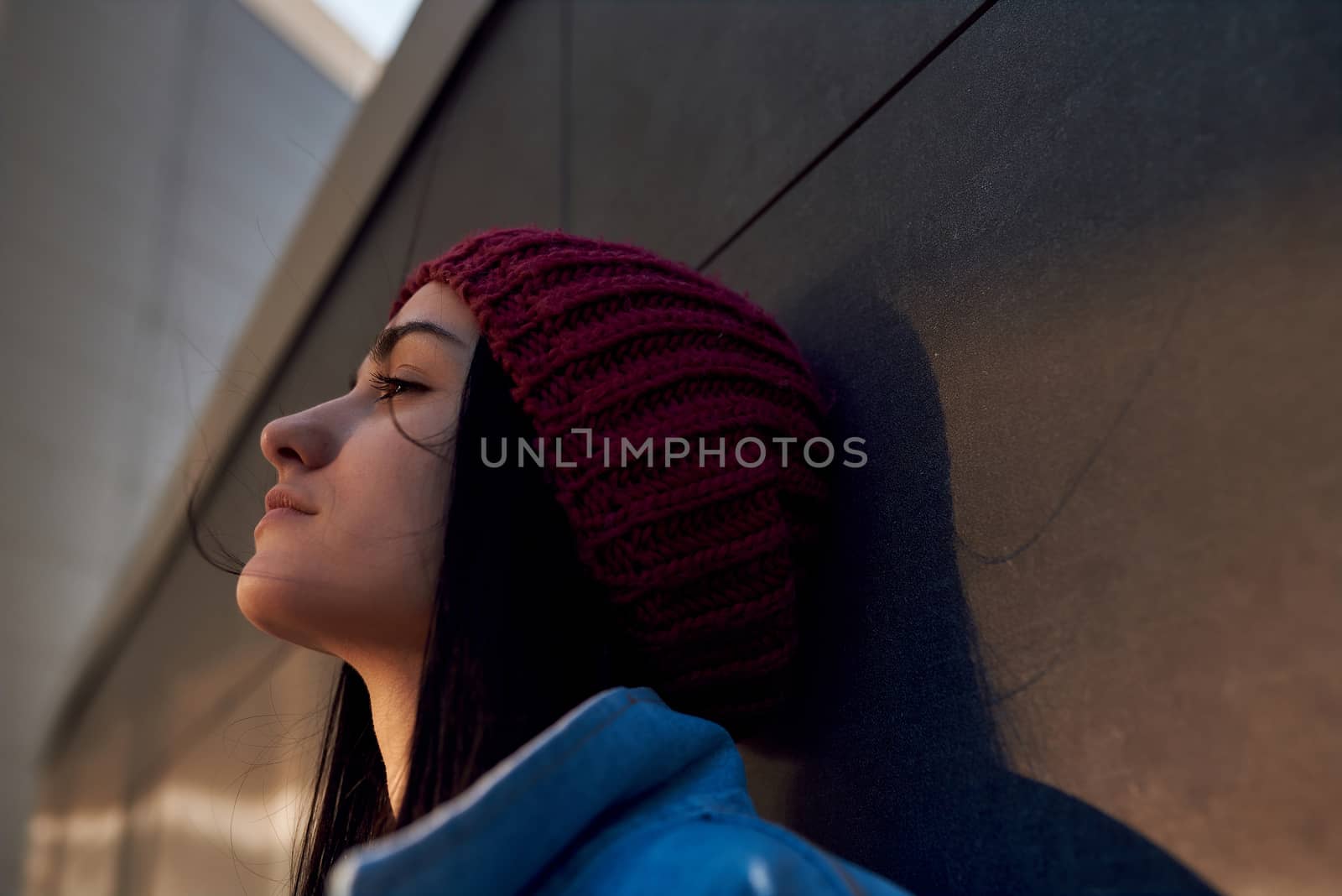 Close up portrait of a pretty female brunette in a knitted cap and denim coat thinking about something on urban background
