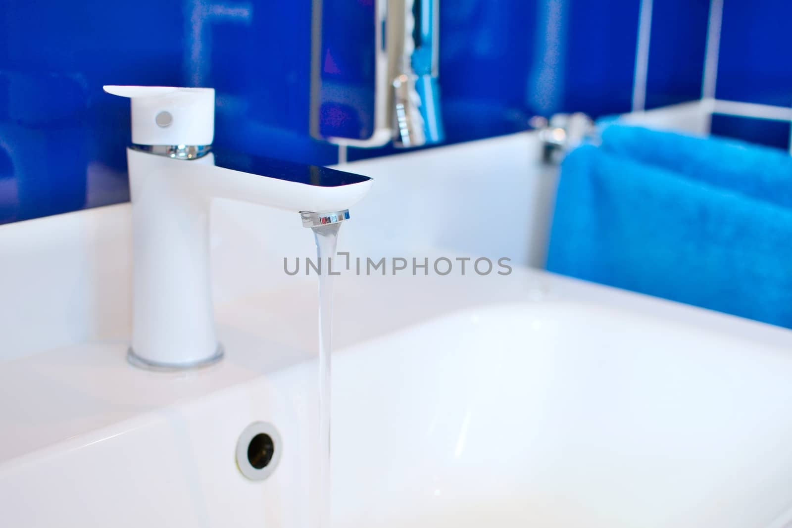 White sink faucet closeup with flowing water in bathroom with blue tiles.