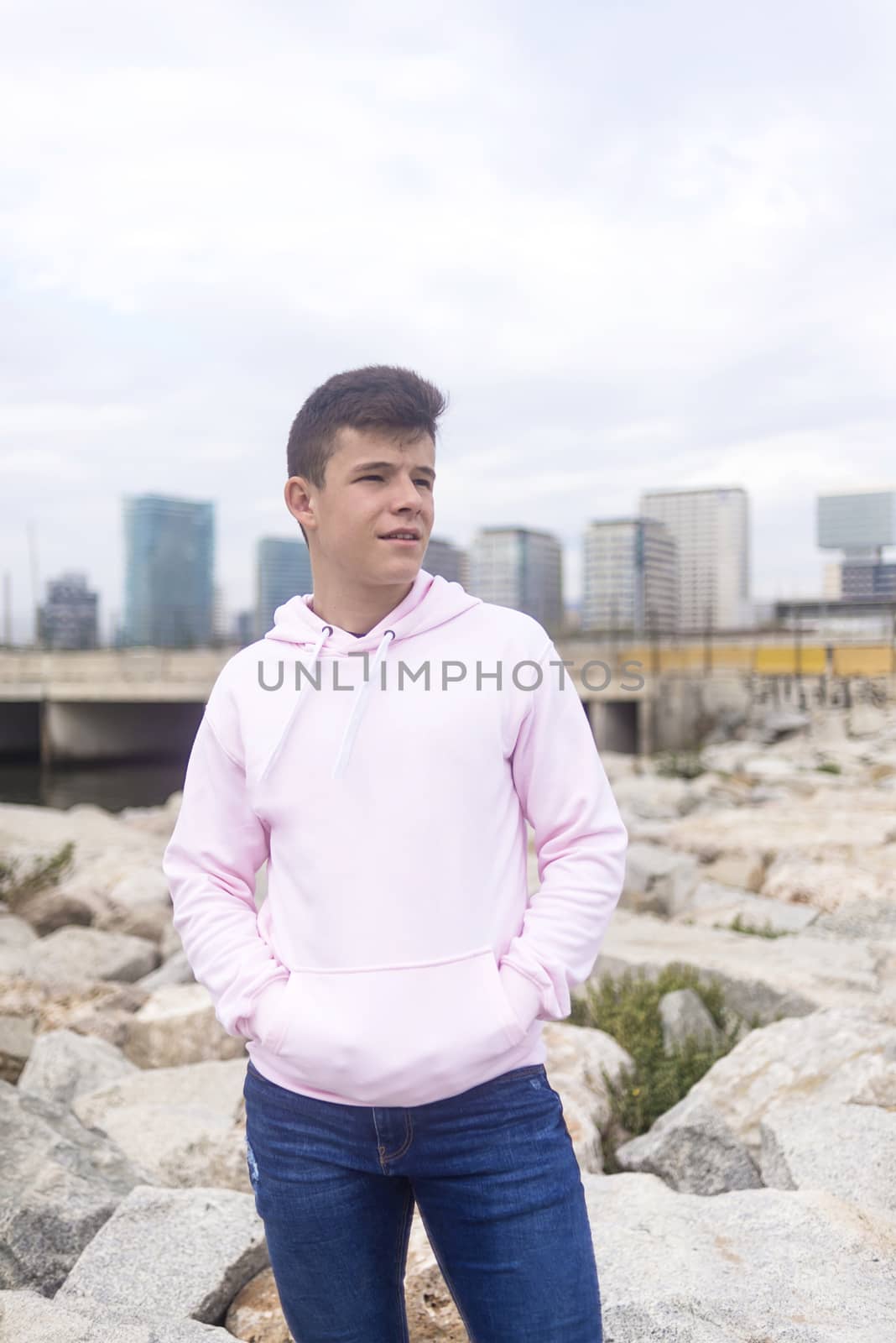 Young teenager male standing against cityscape with hands on pocket on breakwaters while looking away