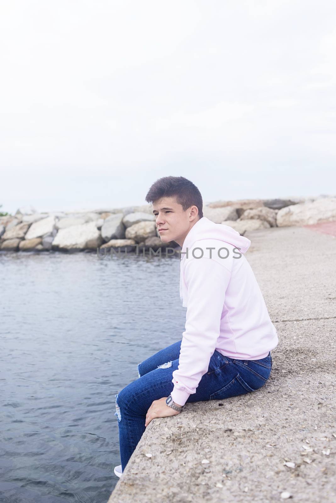 Young pensive teenager male sitting on breakwaters while looking at camera by raferto1973