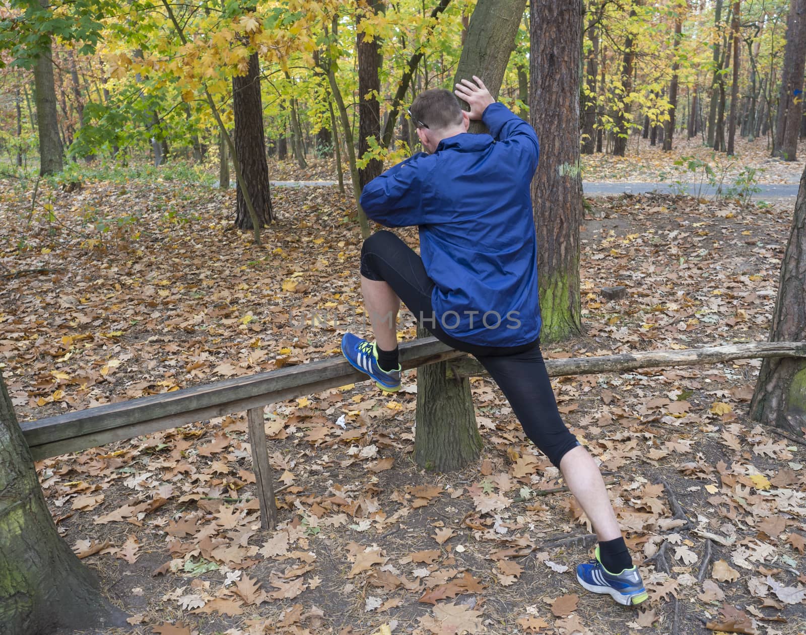 Young athlete runner man stretching his legs in blue jacket and shoes in the autumn woods, warming up ready for jogging on the forest road outside. Fitness and sport concept. by Henkeova