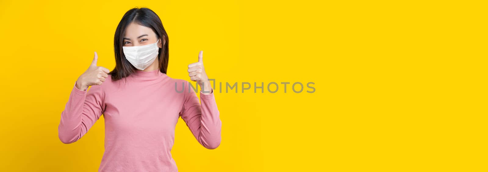 Asian smiling woman wearing mask and thumb up action to be grad, banner or showing on isolated yellow color background, Coronavirus pandemic, covid19 outbreak, banner and beauty with covid19 protect