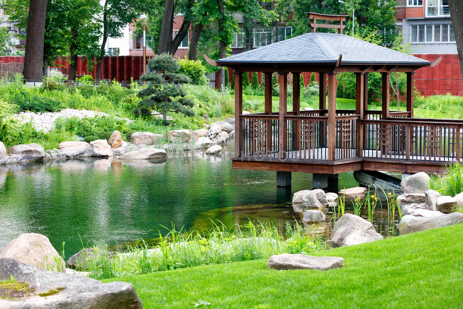 A wooden openwork gazebo on stilts stands in the middle of a forest lake, surrounded by a sheared green lawn with stone boulders on the edges under soft sunlight.