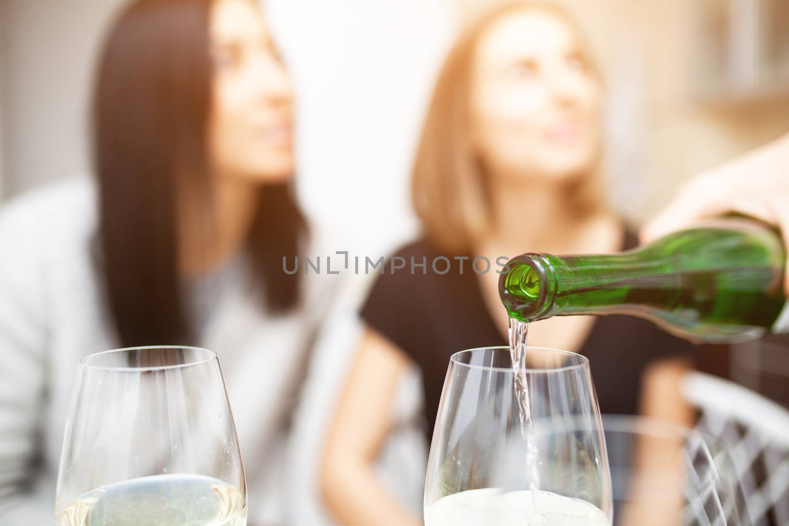 A male hand in the foreground pours wine into glasses. There are two female figures in the background. Friends meeting. Two unrecognizable girls drink wine in the home kitchen.