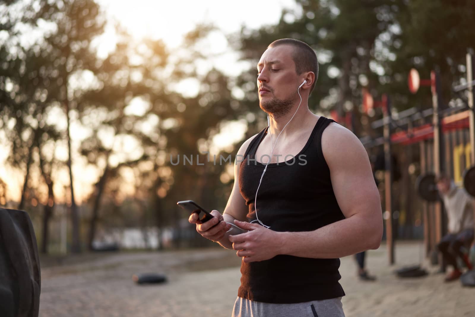 Young adult caucasian athlete listening phone music white headphones after workout. Handsome sportsman resting after cross training exercises sunset background. Healthy lifestyle concept.