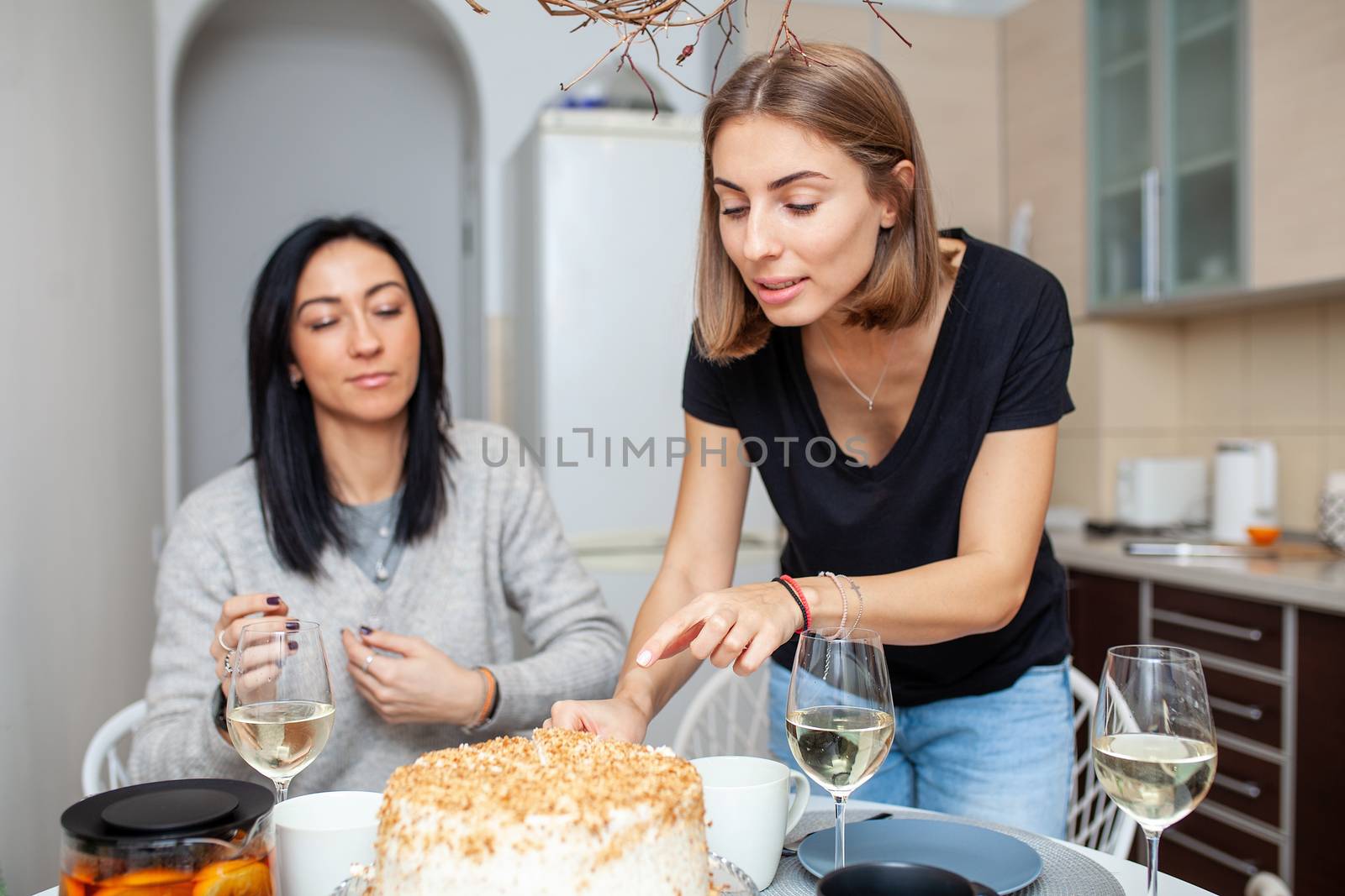 Friends meeting with wine and cake in the modern style kitchen. Women smile and joke by andreonegin