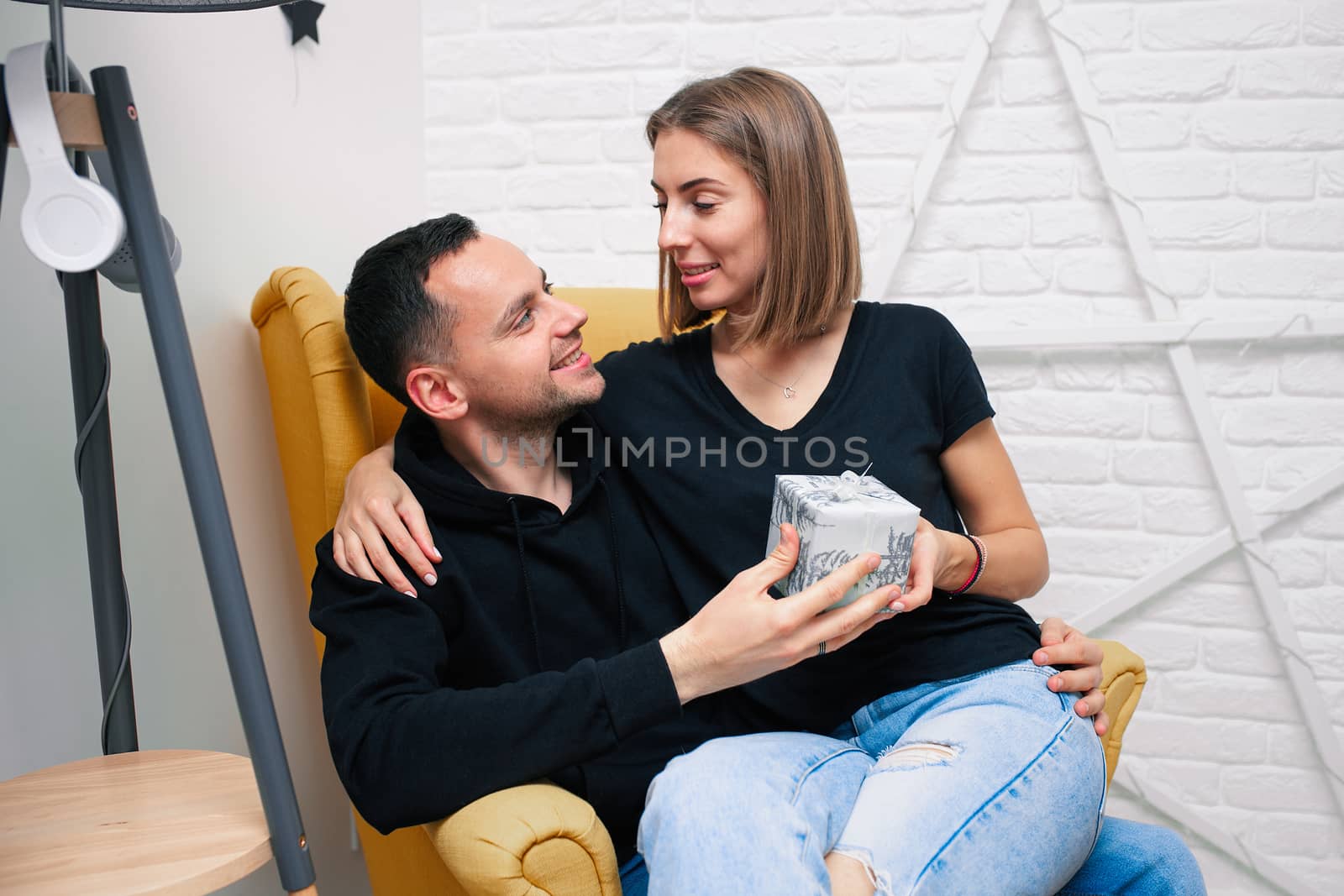 Portrait of couple young in a cozy room. A man sits on a yellow chair. A woman sits on the lap of a man and holds a gift Christmas family holidays. Modern style interior.