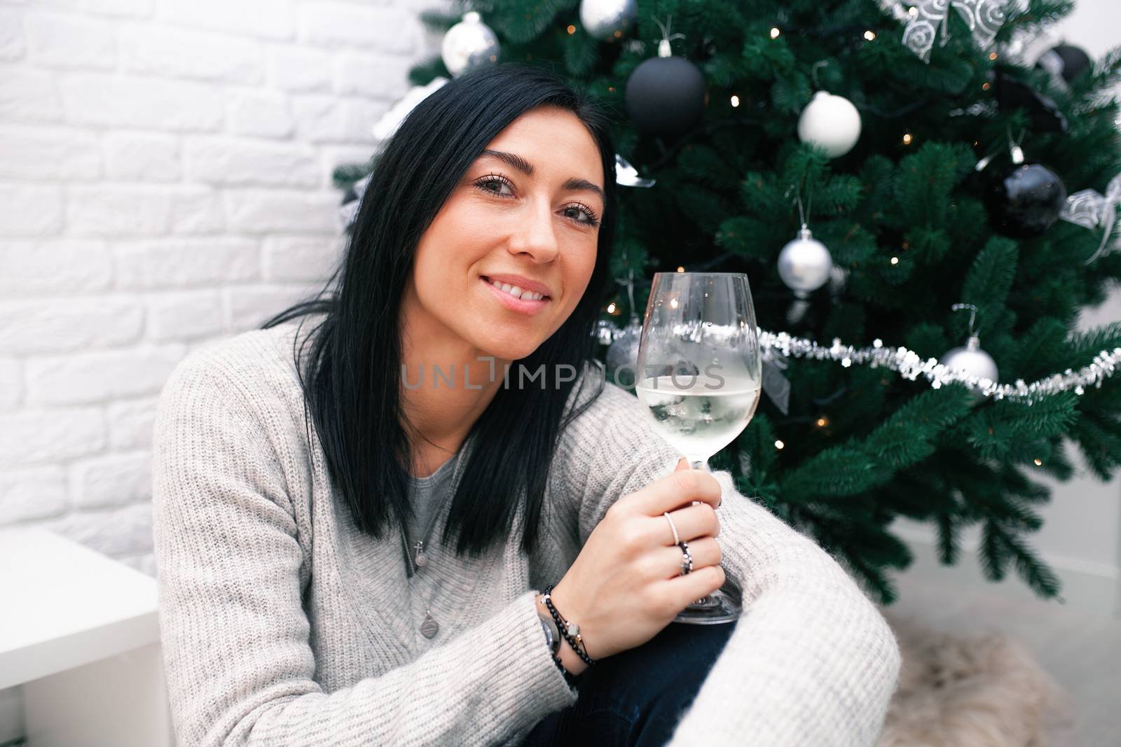 A young woman sits near a Christmas tree, holding a glass with white wine and a Christmas toy. Preparation for the New Year's celebration.