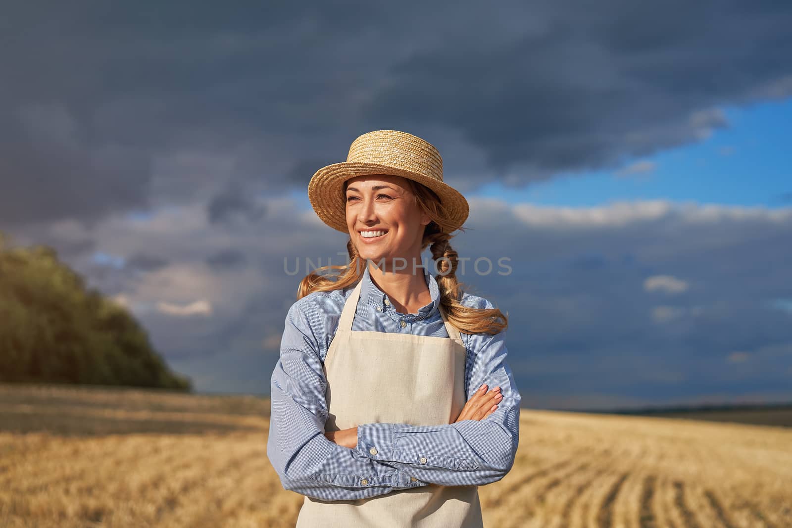 Woman farmer straw hat apron standing farmland smiling Female agronomist specialist farming agribusiness Positive caucasian worker agricultural field arms crossed cloudy sky background before rain