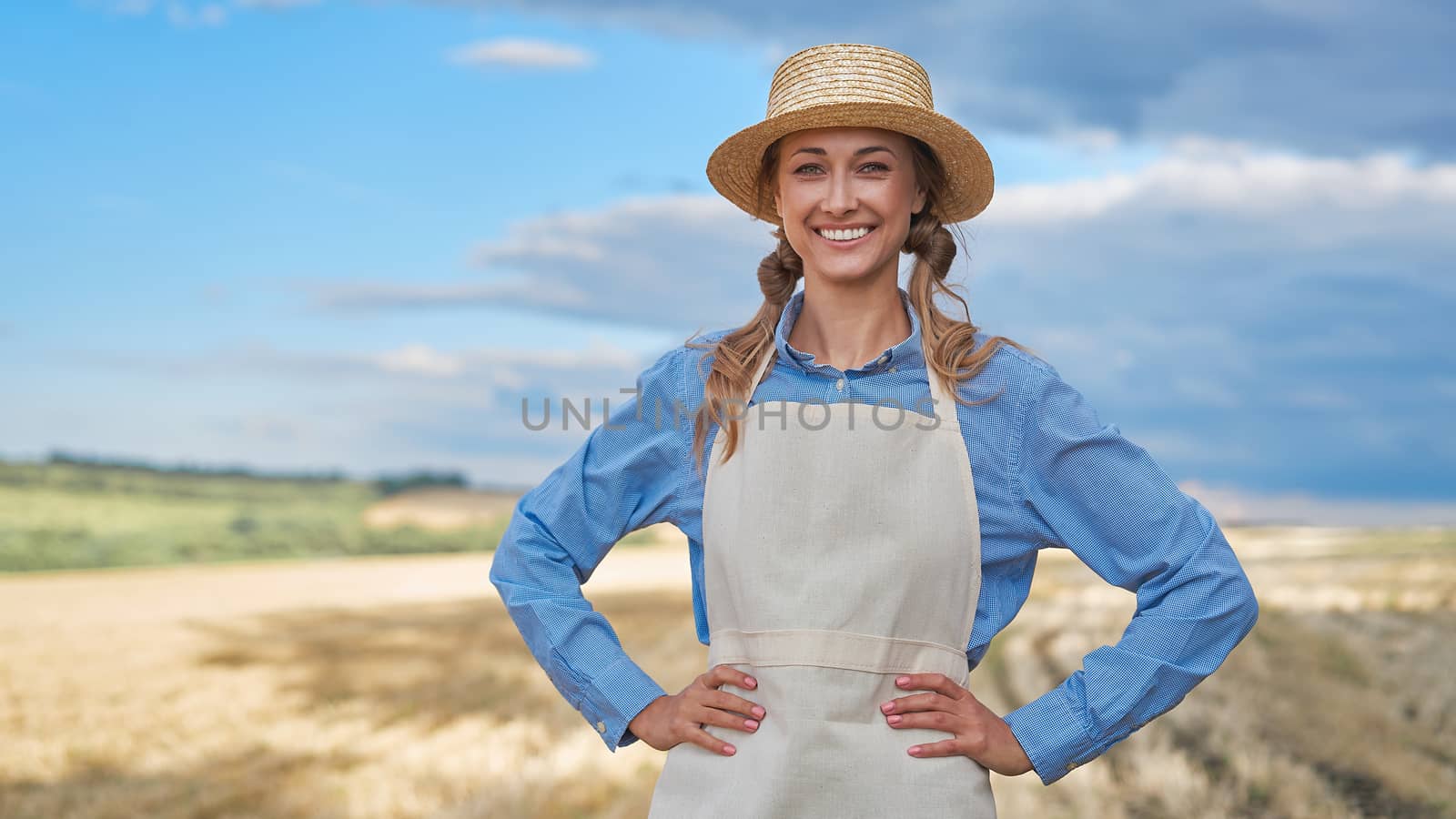 Woman farmer straw hat apron standing farmland smiling Female agronomist specialist farming agribusiness Happy positive caucasian worker agricultural field Girl hands on waist cloudy sky background