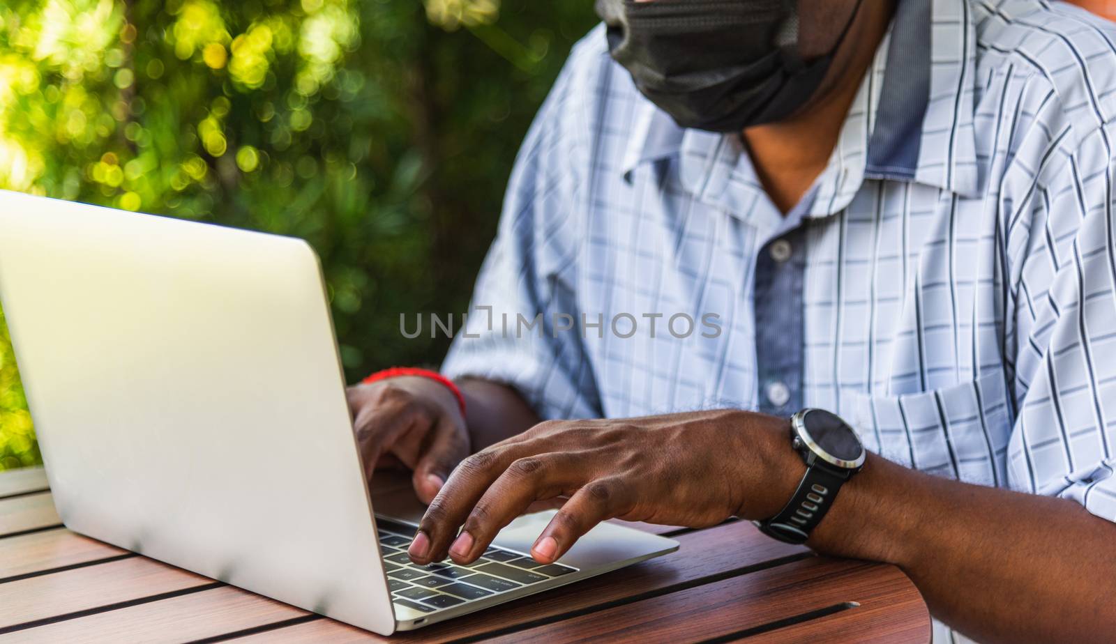 Happy Asian black businessman person or office worker wear face mask protect sitting on desk work from home using laptop computer typing keyboard and connect networking online at the coffee cafe shop