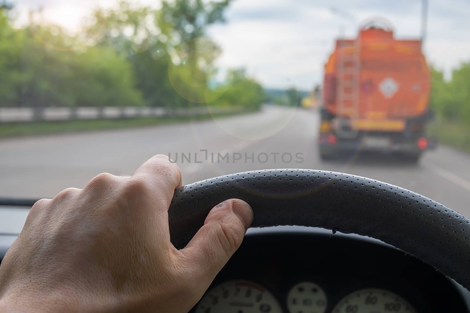 view of the driver hand at the wheel of a car that is driving behind a fuel truck on the road