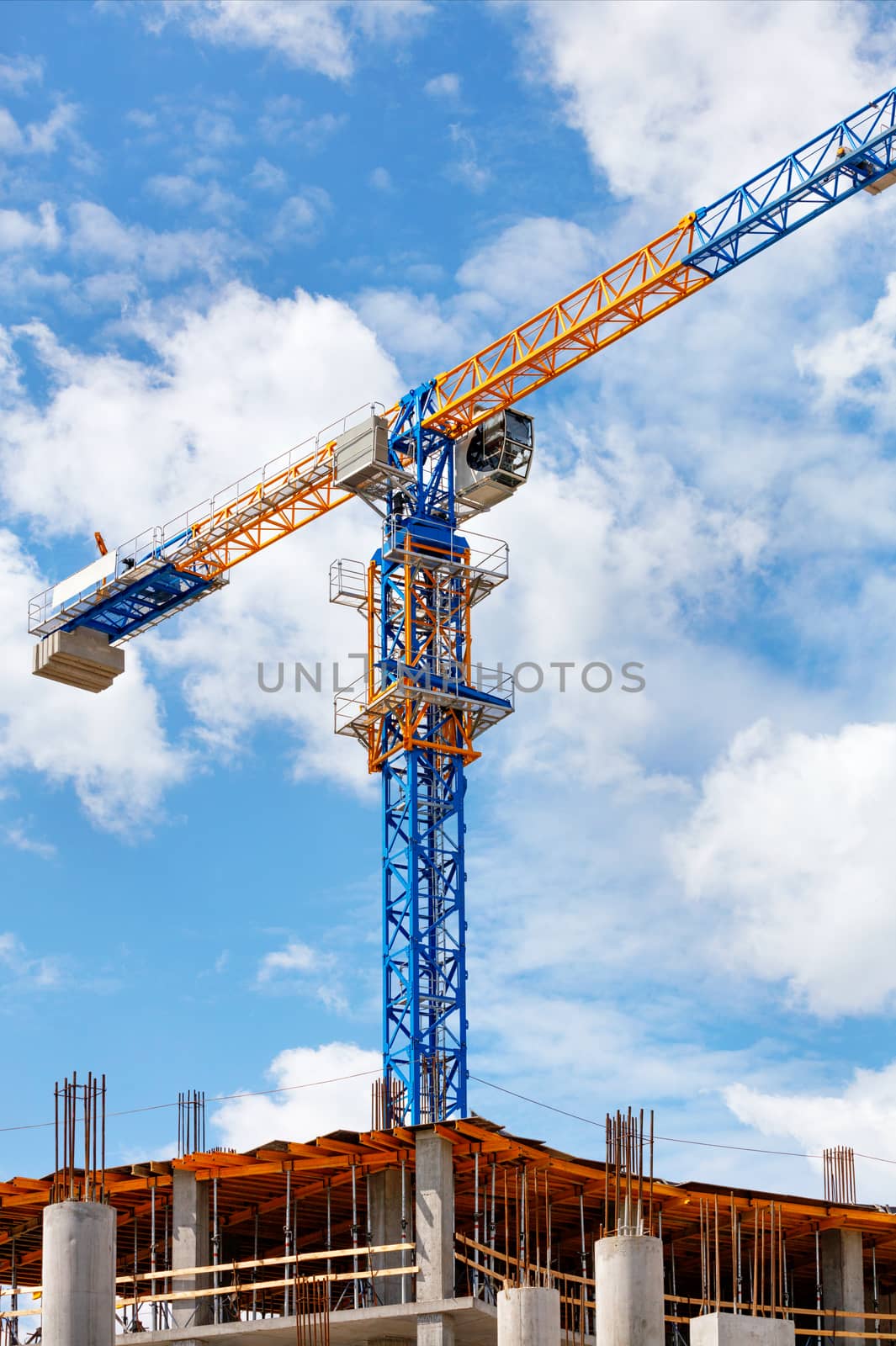 The arrow of a yellow and blue high-rise tower crane rises above the construction site diagonally against a background of blue sky and white clouds, copy space.