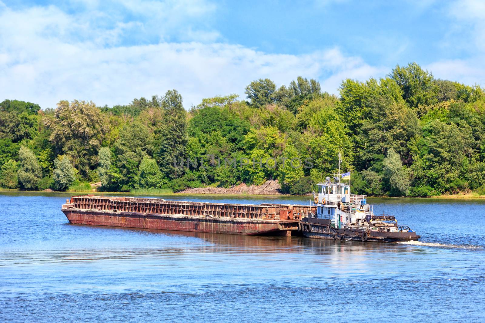 River tugboat pushes an empty rusty barge along the river against the background of coastal greenery and calm surface of the water, the concept of river freight transportation, copy space.
