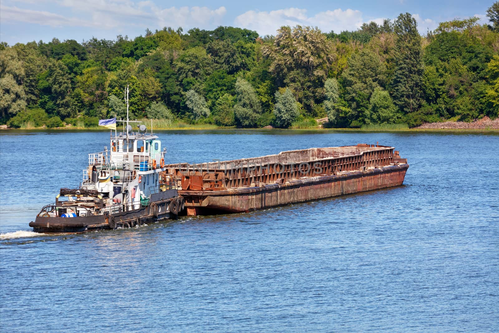 A river tugboat with an empty rusty barge goes crosses a wide river. by Sergii