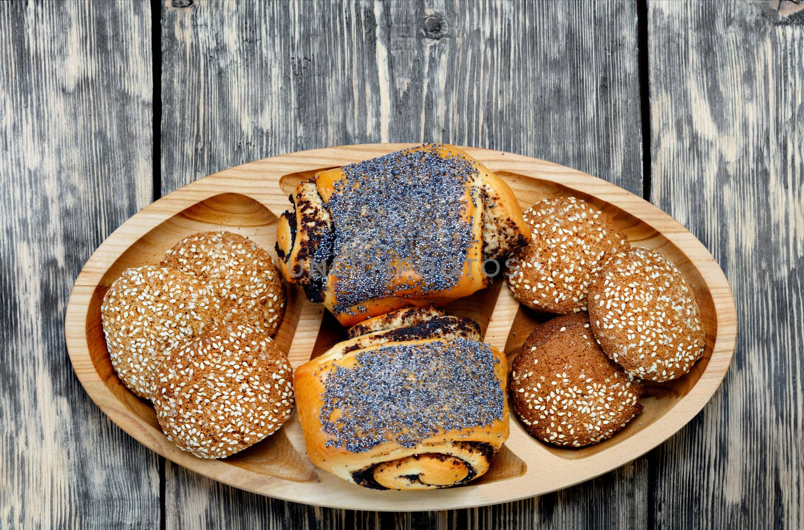 Butter appetizing buns with poppy seeds and sesame cookies on a wooden oval board on an old wooden table, rustic style, copy space.