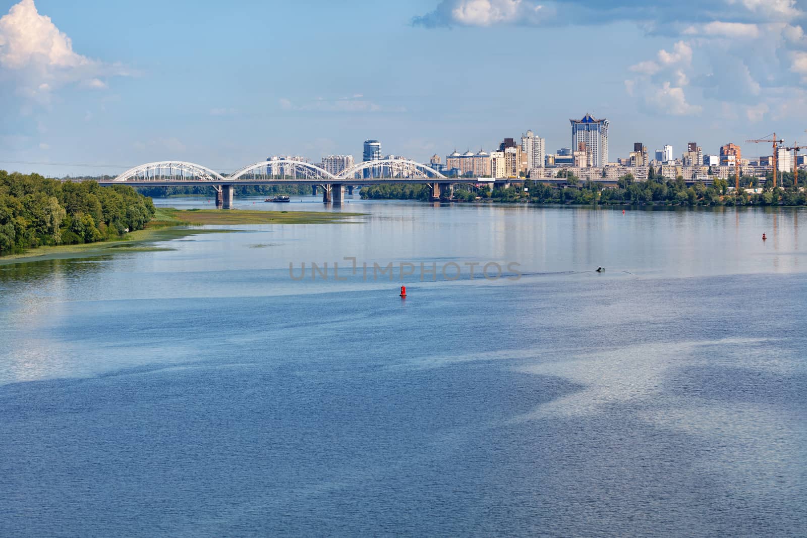 The water surface of the wide Dnipro River in the foreground, immersed in the green of the banks, a white railway bridge and new residential areas of Kyiv on the horizon, copy space.
