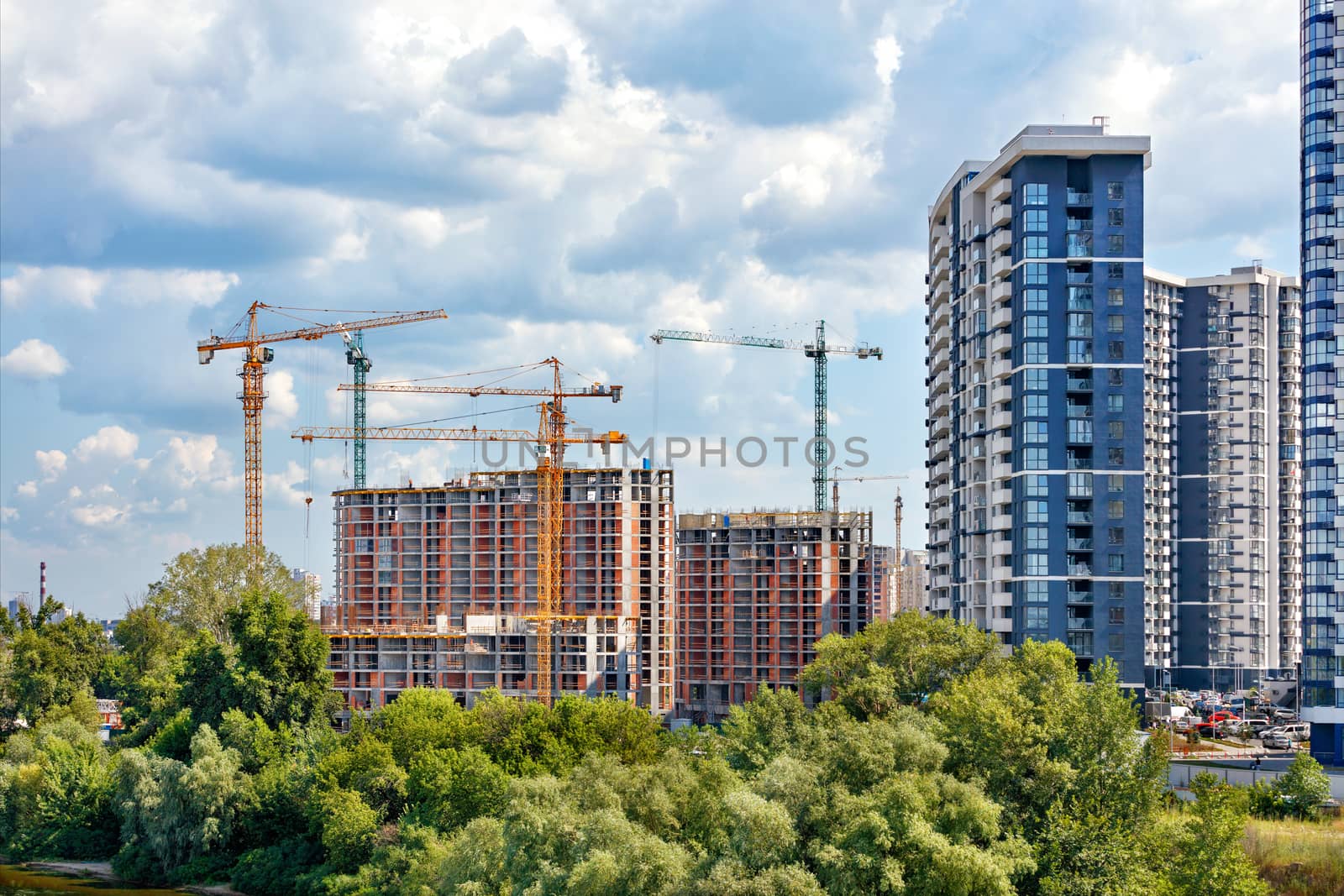 Construction site of a multi-storey residential complex on the river bank against the background of coastal trees, cloudy sky and facades of new buildings, copy space.