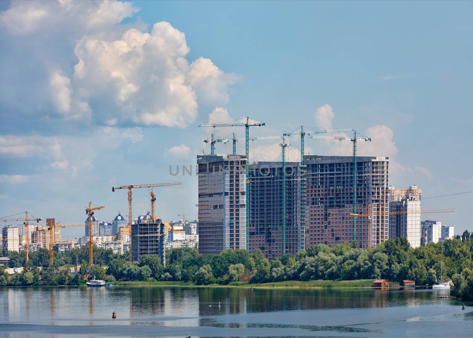 Construction of modern residential high-rise buildings along the river bank of the Dnipro, tower cranes rush upward touching the clouds. by Sergii