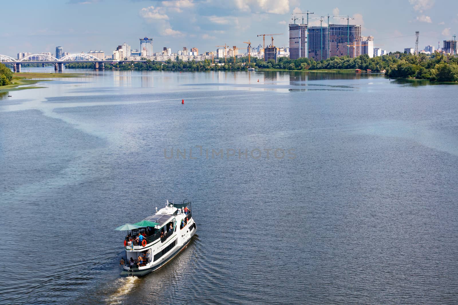 A river tram with tourists resting goes along the river against the backdrop of coastal greenery and high-rise buildings on the horizon on a warm summer day. River passenger transport concept, copy space.