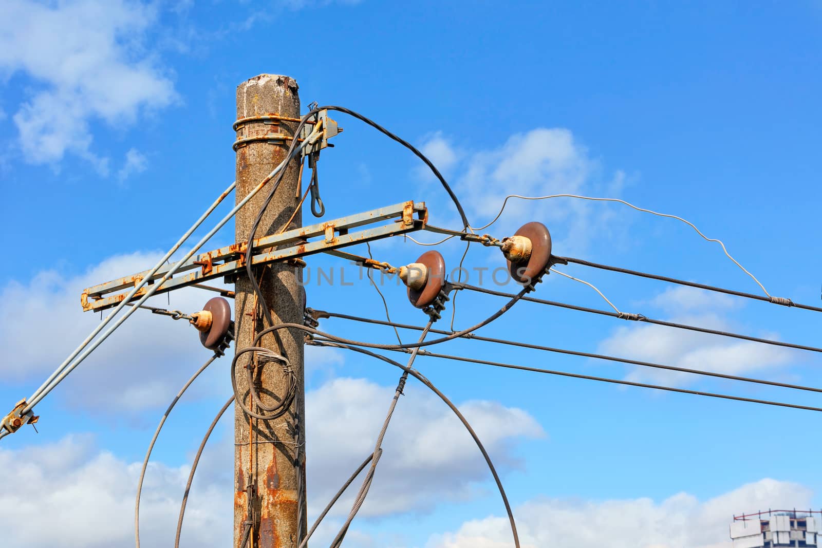 The top of an old concrete pillar with electric wires and a connected three-phase current stands against a blue cloudy sky.
