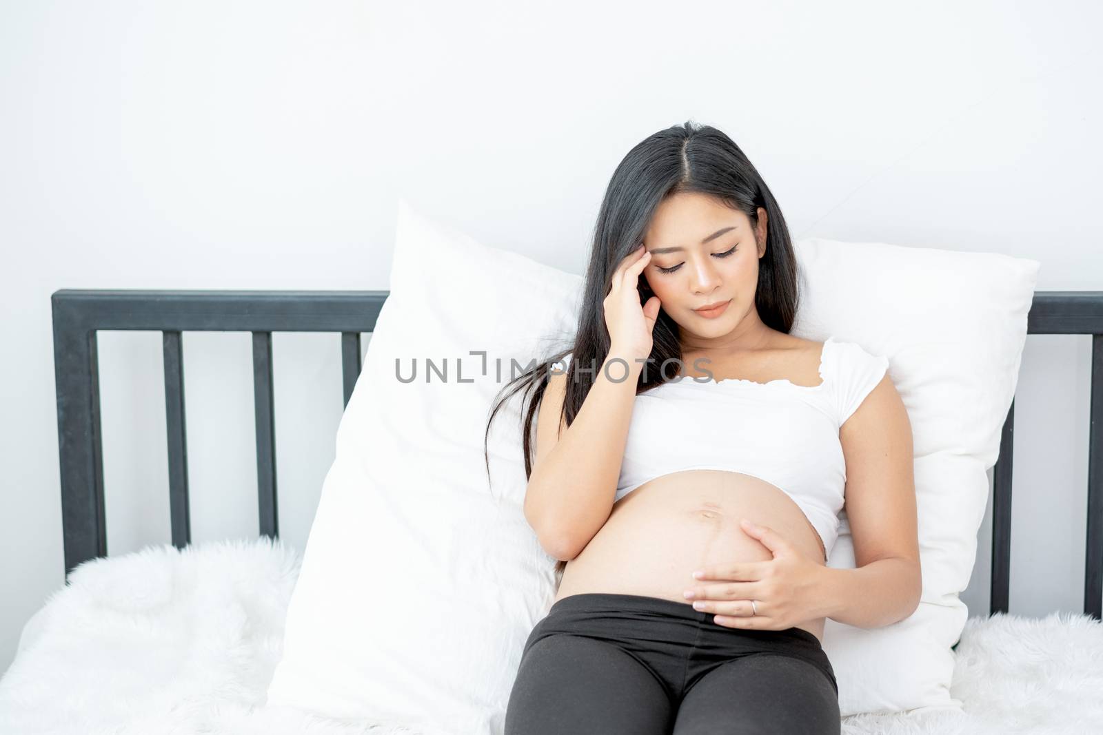 Beautiful Asian pregnant woman show action of stress and headache lie on bed with morning light. Concept of sickness of mother during pregnancy and need to take care and support at home.