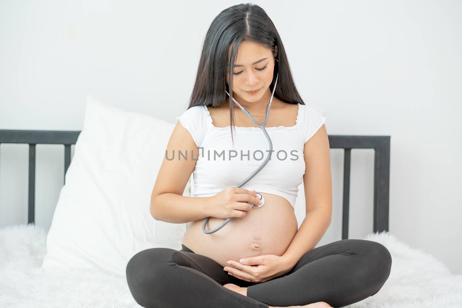 Portrait of Asian pregnant mother use stethoscope to listen sound of baby in her womb during sit on bed with morning light. Concept of good take care baby in the womb of healthy mother stay at home.