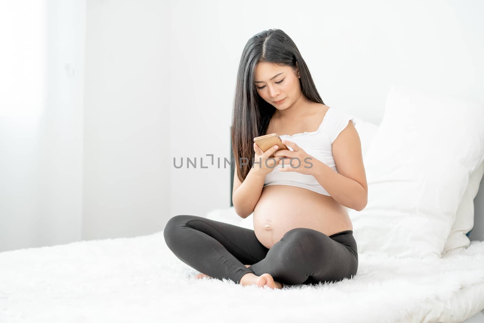 Portrait of beautiful Asian pregnant woman sit on bed and use mobile phone with morning light. Concept of new normal lifestyle to stay safe at home.