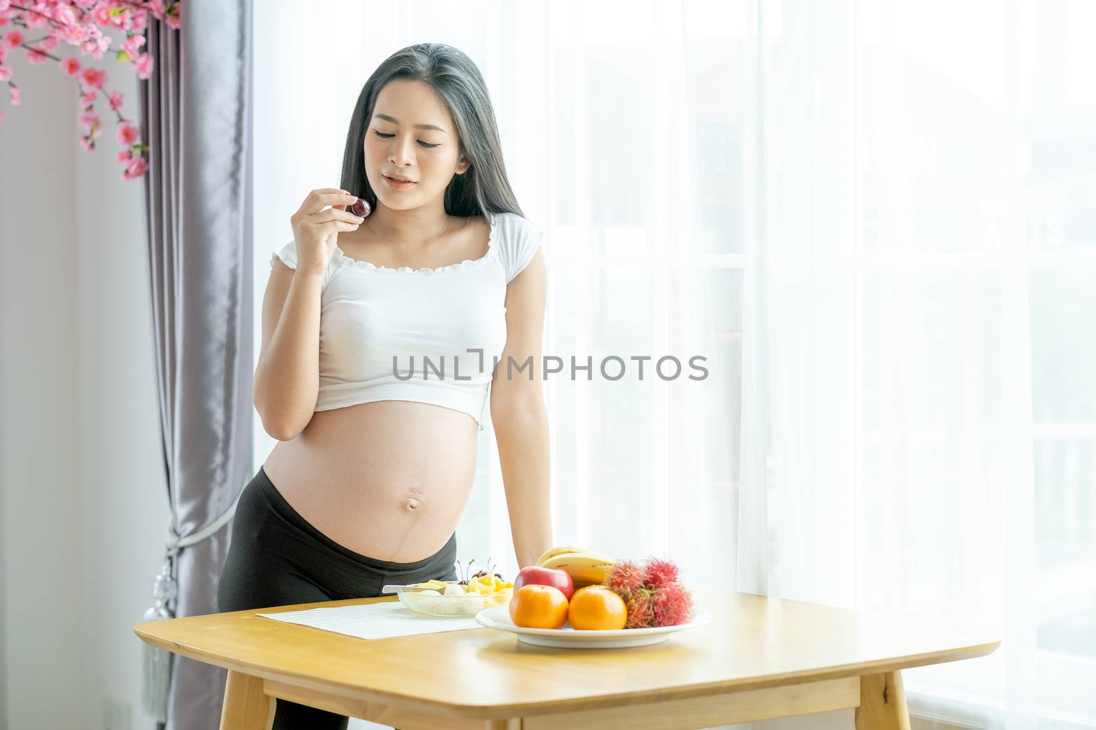 Soft blur of beautiful pregnant woman hold berry and stand near table with various types of fruits in front of white curtain with morning light. Concept of good and healthy food for pregnant people.