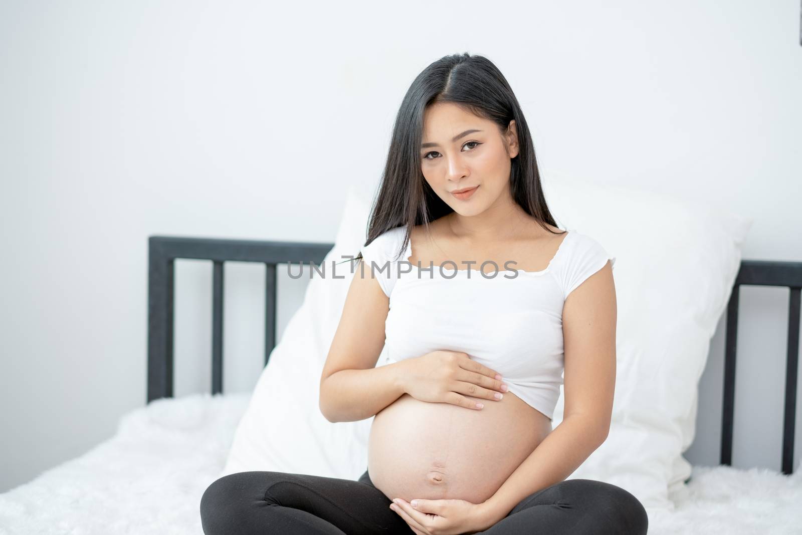 Portrait of beautiful Asian pregnant woman sit on bed and touch and look to her belly also smiling. Concept of good take care baby in the womb of healthy mother and stay at home.