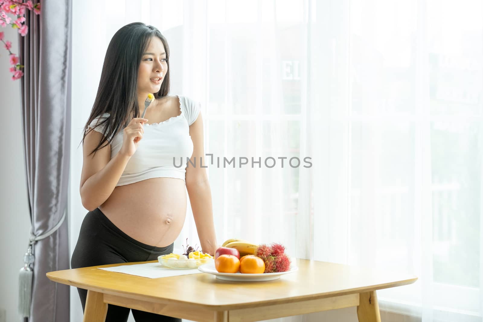 Beautiful pregnant woman hold folk with some fruits and stand near table with various types of fruits in front of curtain with morning light. Concept of good and healthy food for pregnant people.